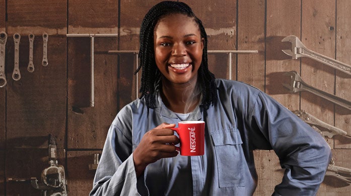 A woman in a blue jumpsuit with a big smile is standing in front of a wooden wall with several workshop tools while holding a red NESCAFÉ mug.​