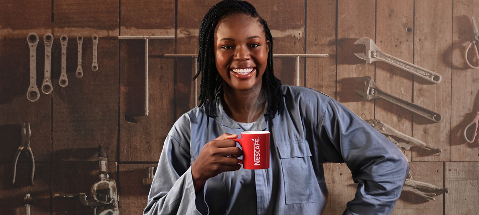 A woman in a blue jumpsuit with a big smile is standing in front of a wooden wall with several workshop tools while holding a